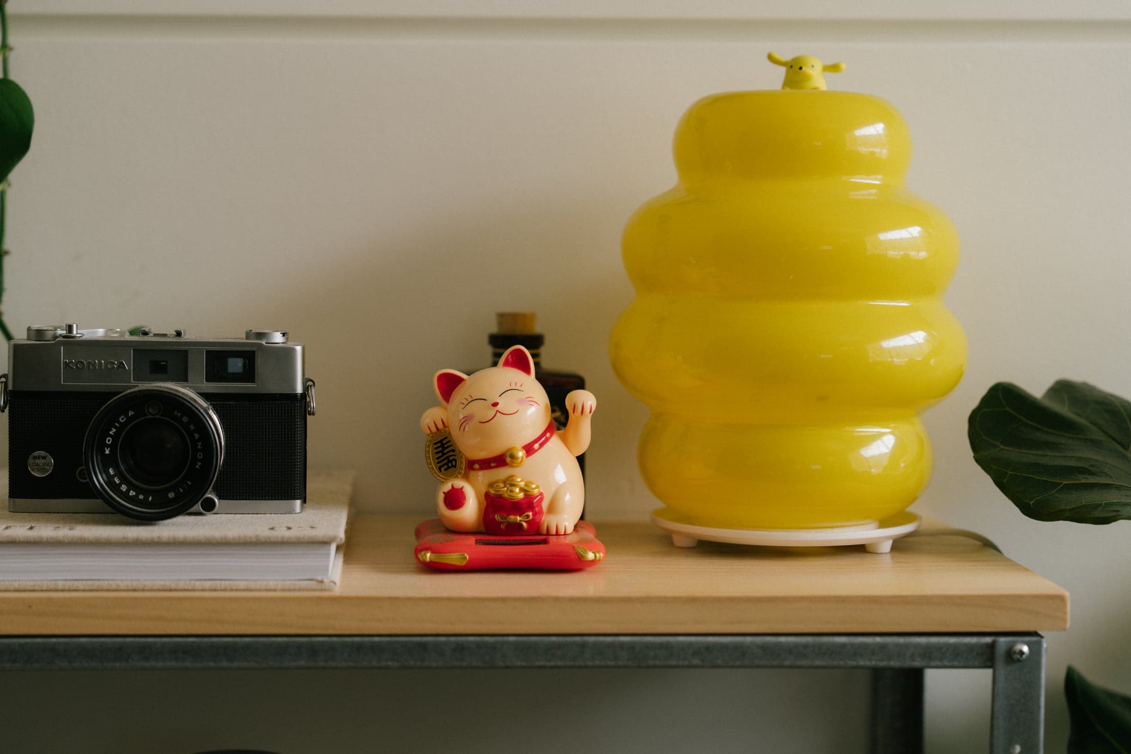 A vintage camera, waving lucky cat and yellow lamp on a studio shelf.