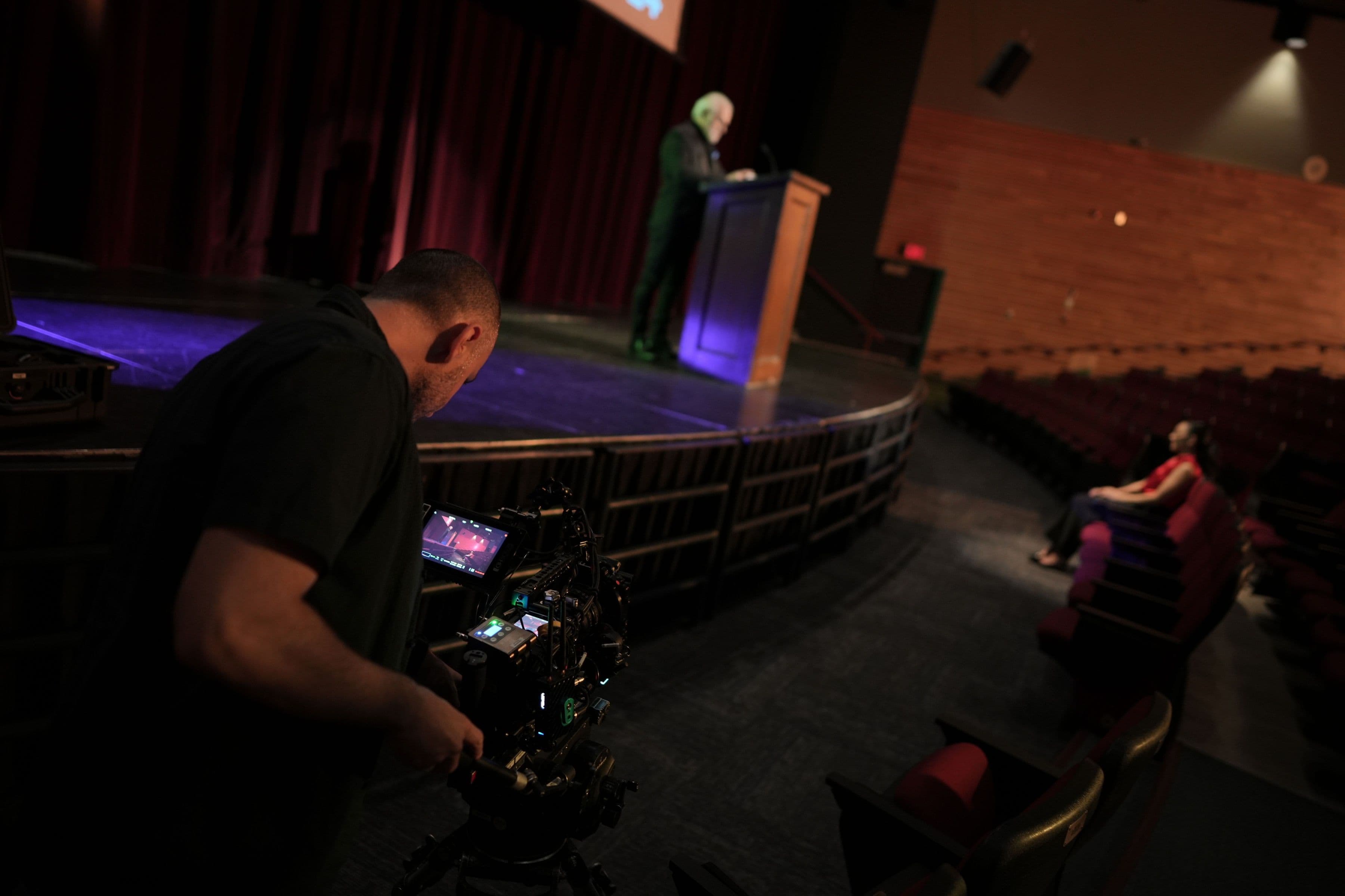 A camera operator filming a speaker at a podium in a theatre.