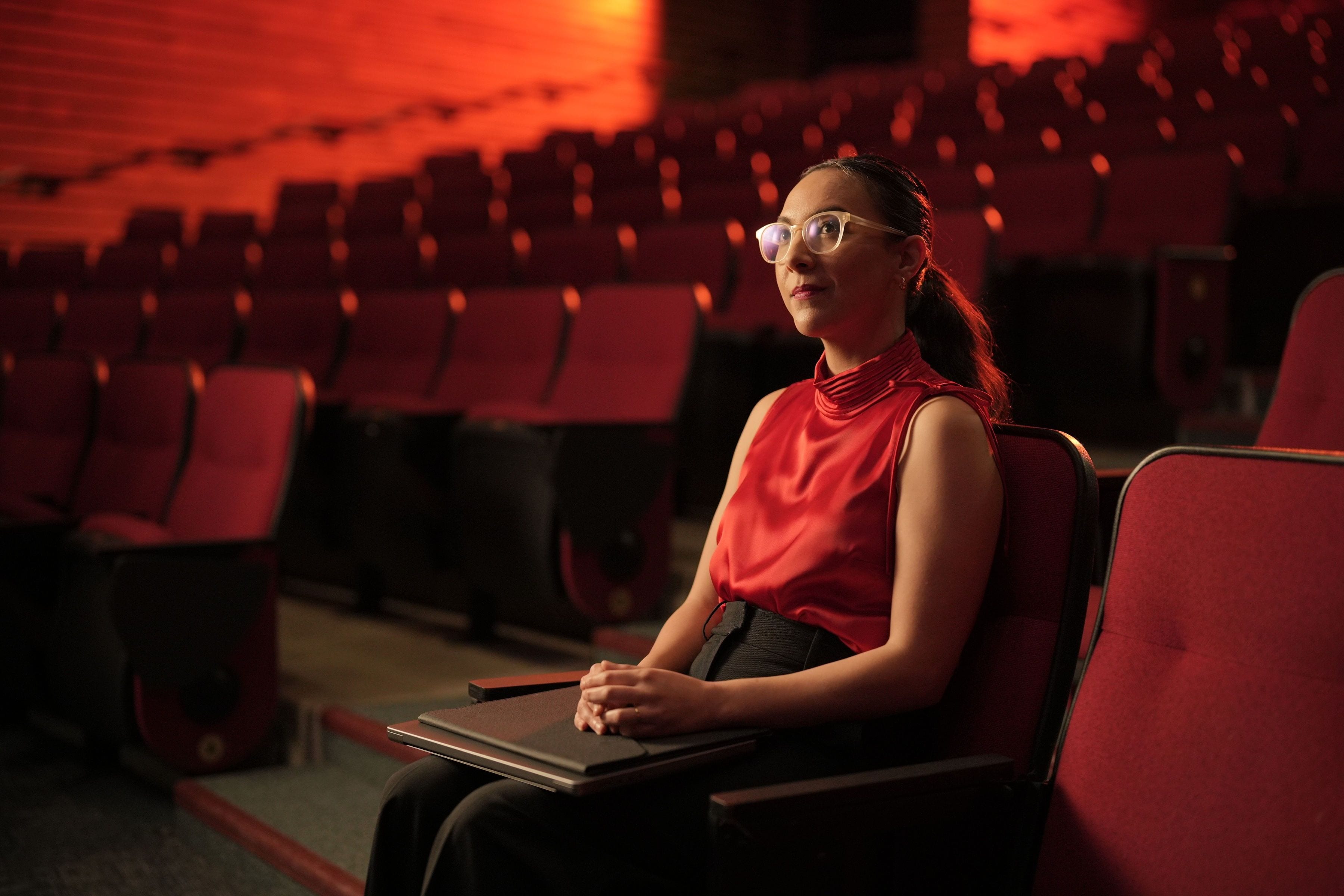 A woman in a red sleeveless top seated among rows of red theatre seats.