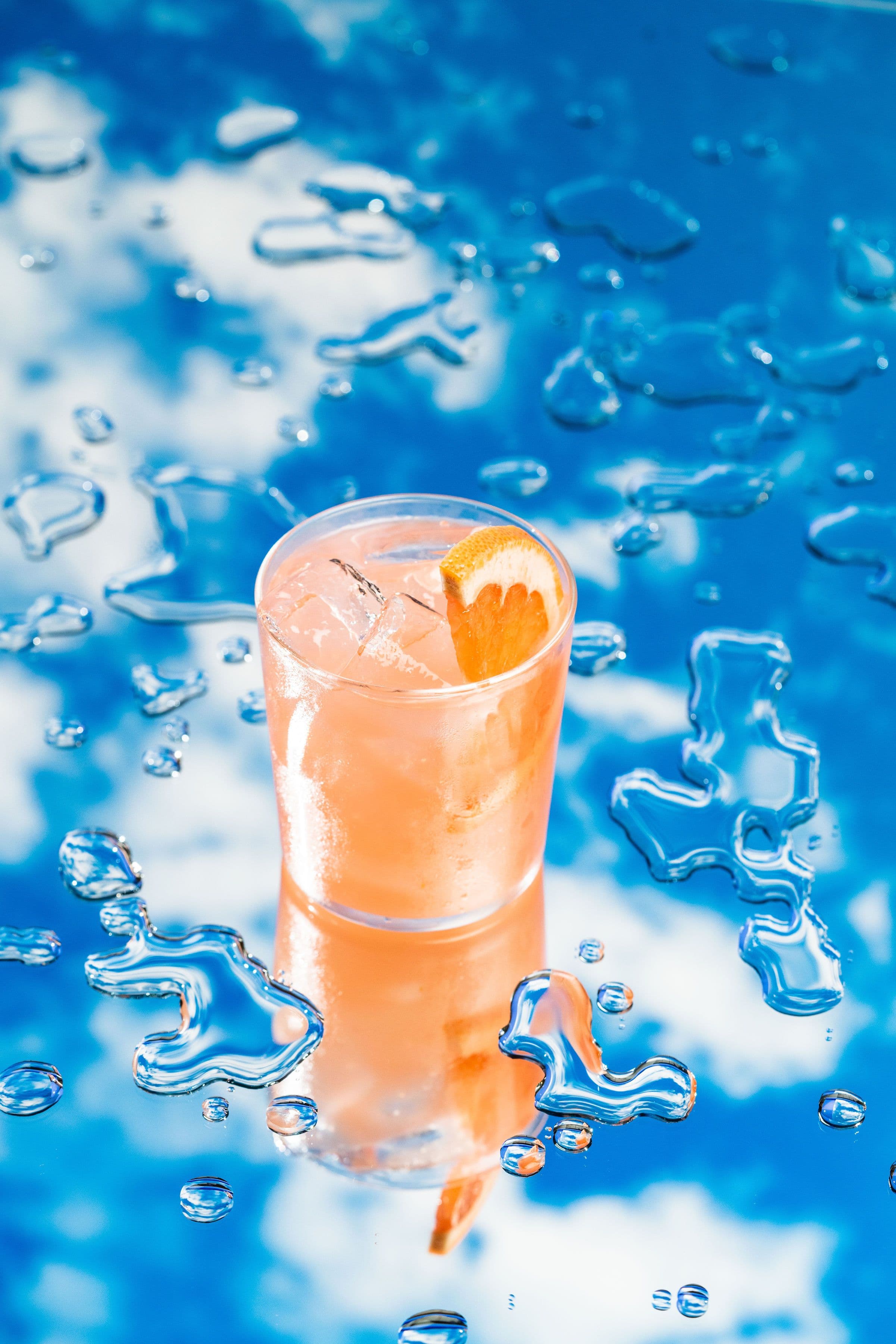 A pale peach cocktail standing on a reflective blue water-inspired set.