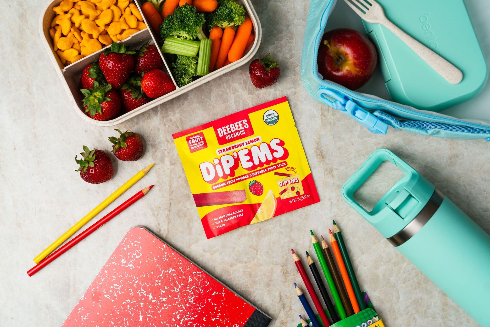 A packet of DeeBee’s Dip’Ems beside a lunchbox of fruit and vegetables, a water bottle and school supplies.