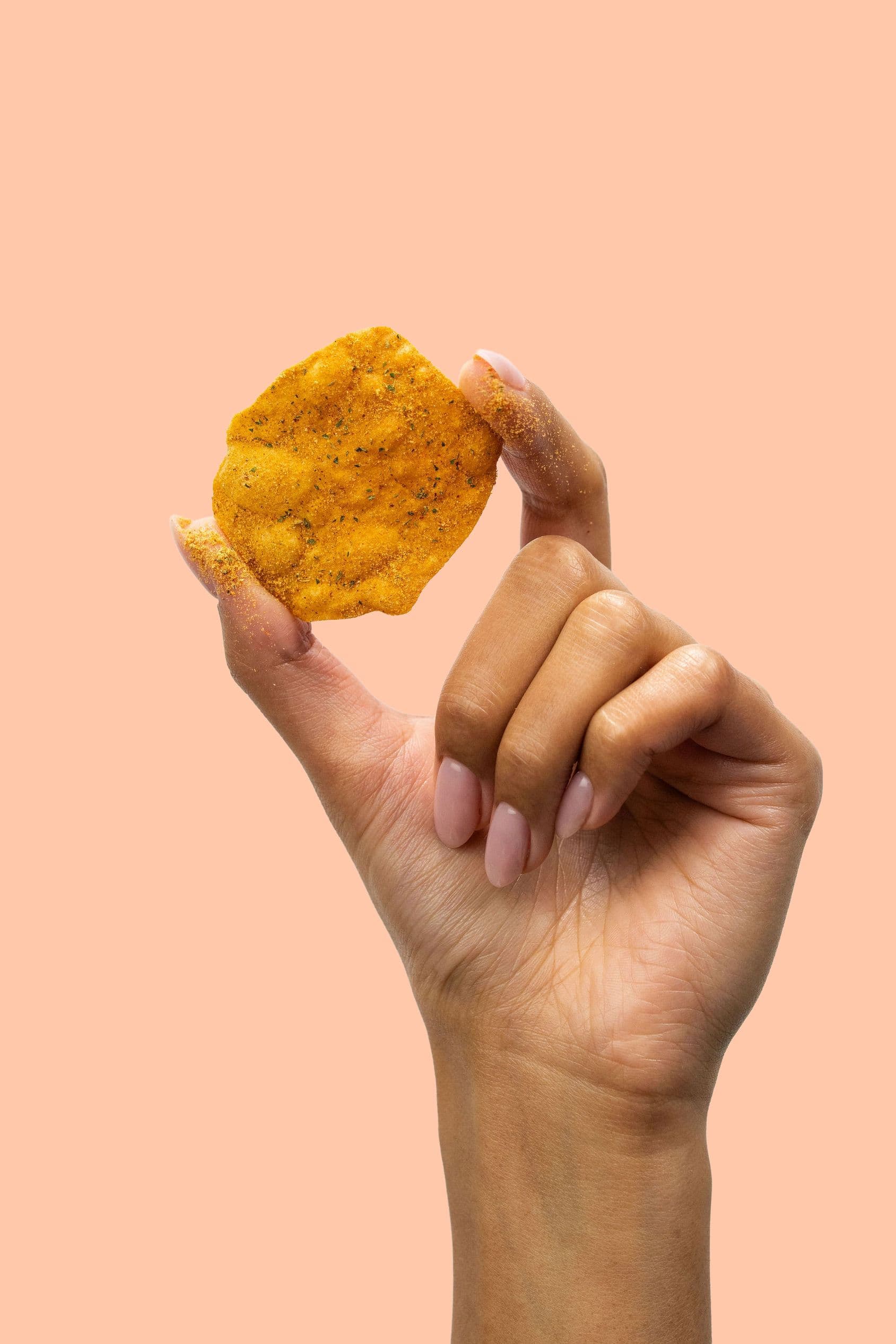 A hand holding a single seasoned potato crisp against a peach background.