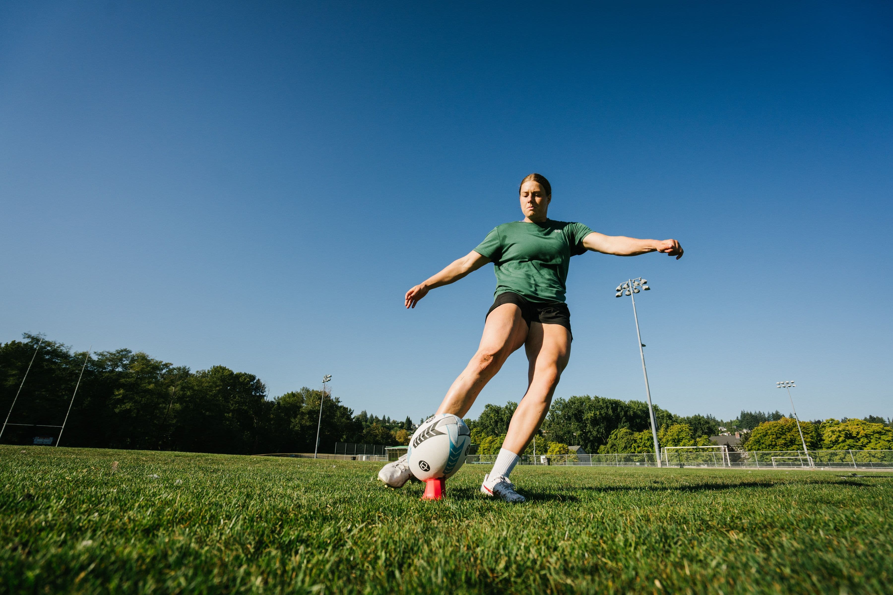 Sophie de Goede kicking a rugby ball from a red tee on a sunny grass field.