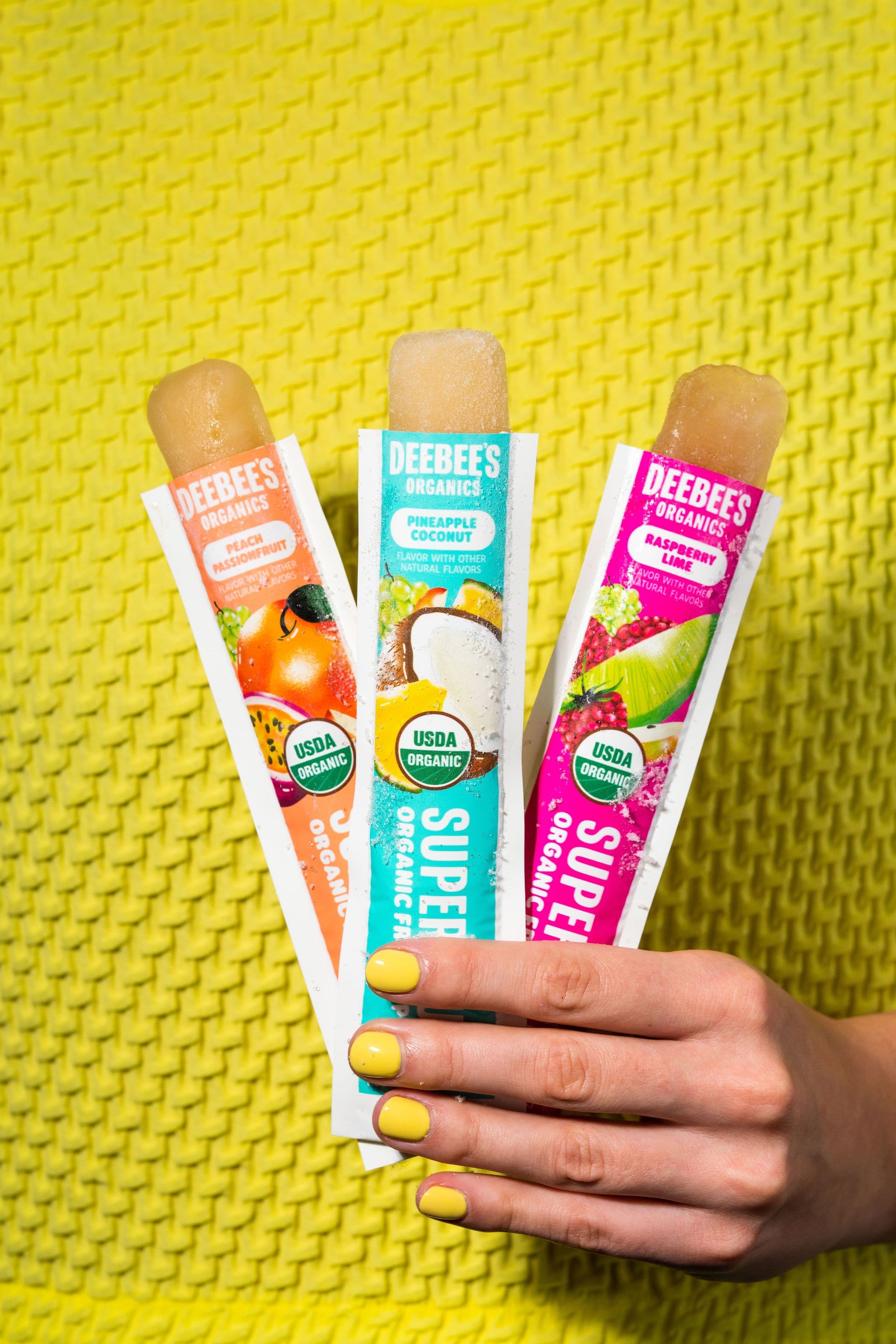 A hand holding three opened DeeBee’s SuperFruit freezies against a bright yellow knit sweater.