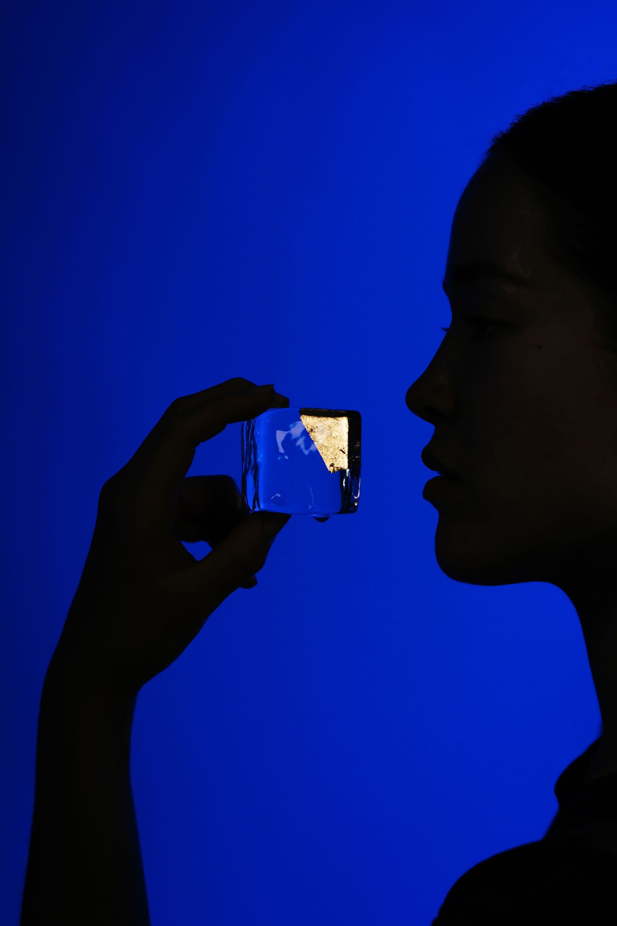 A silhouetted model holds a small clear ice cube with gold leaf near her mouth against a blue background.