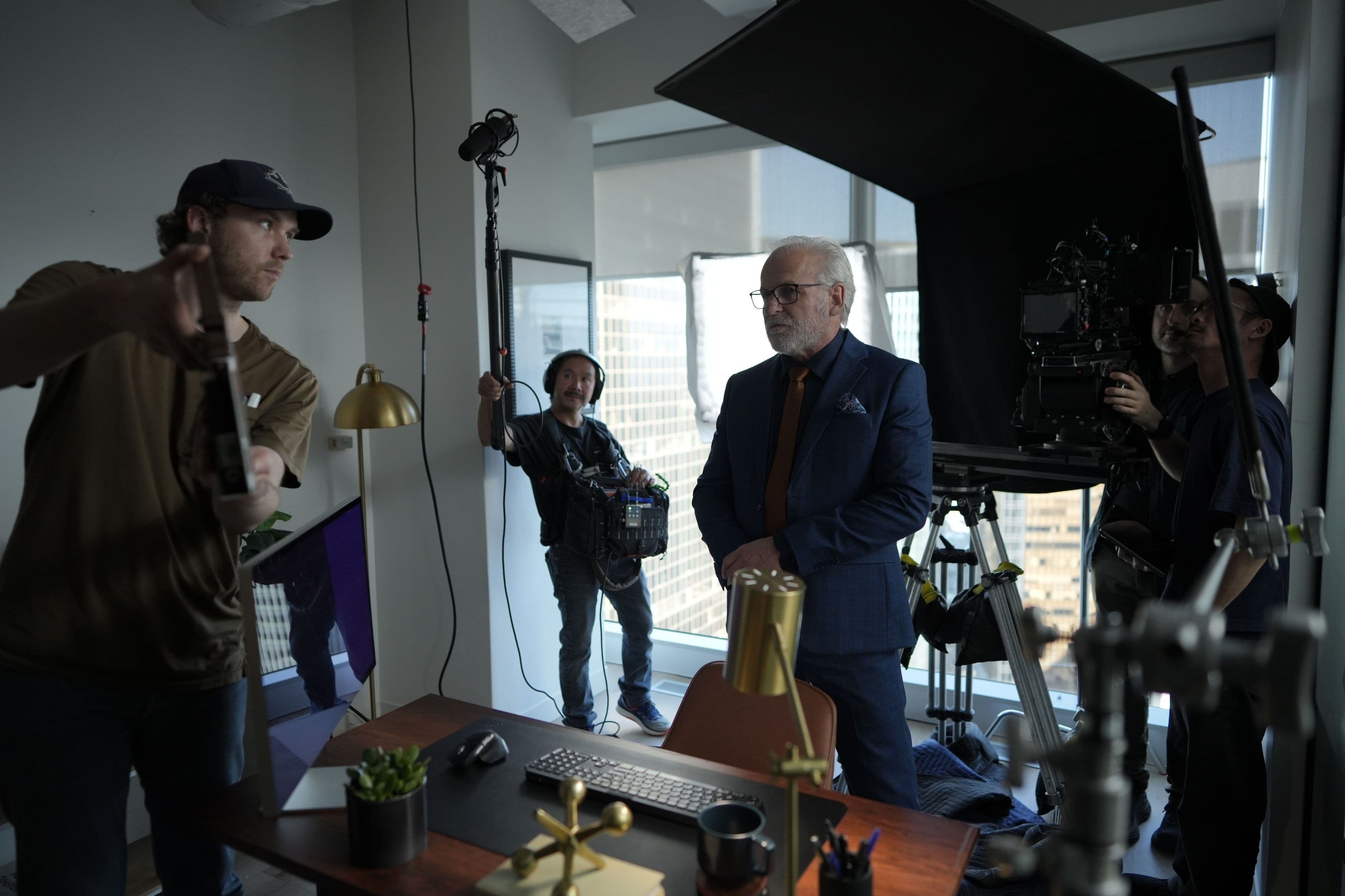 A film crew preparing an office scene with an actor beside a desk and a large overhead light.