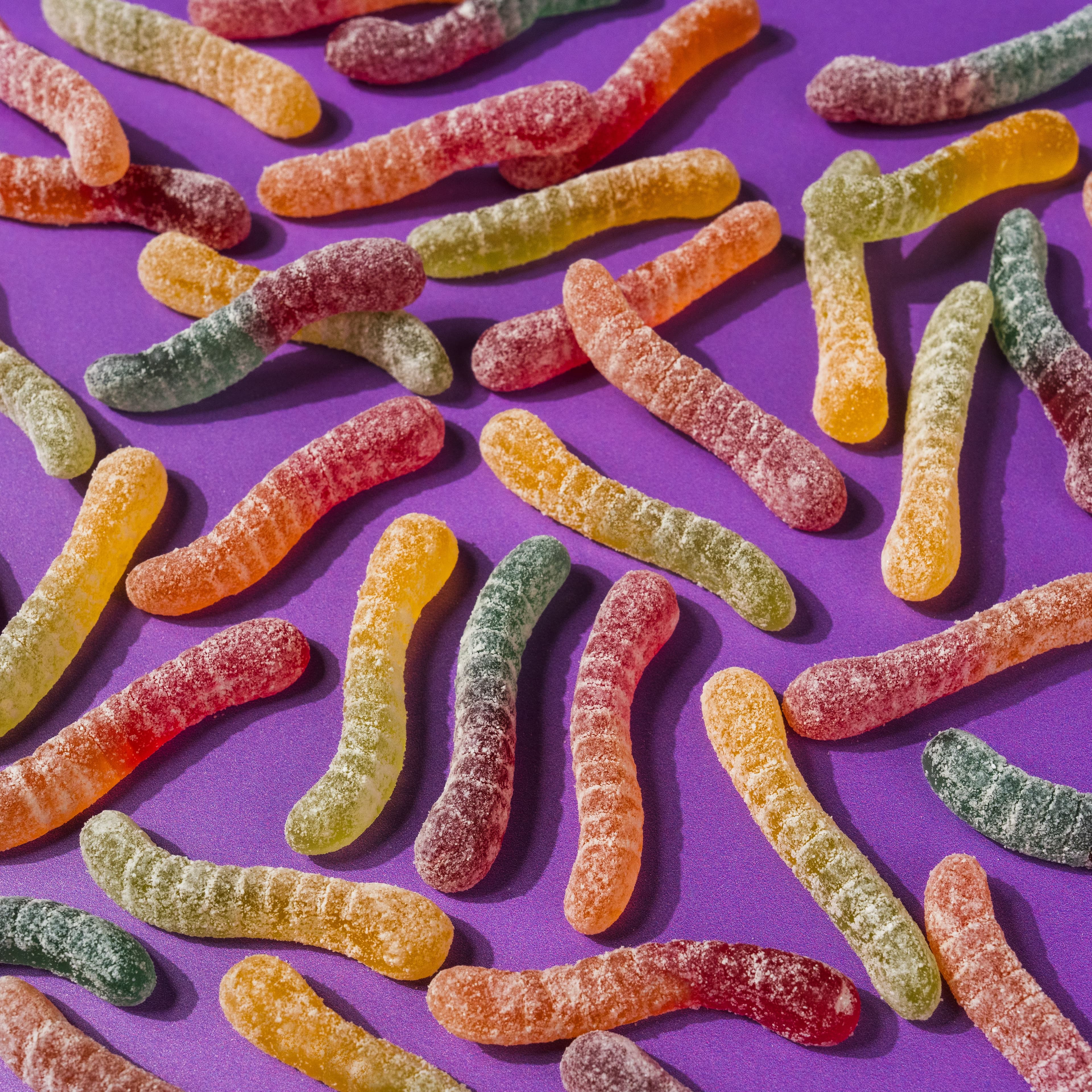 Colourful sour gummy worms scattered across a saturated purple surface.