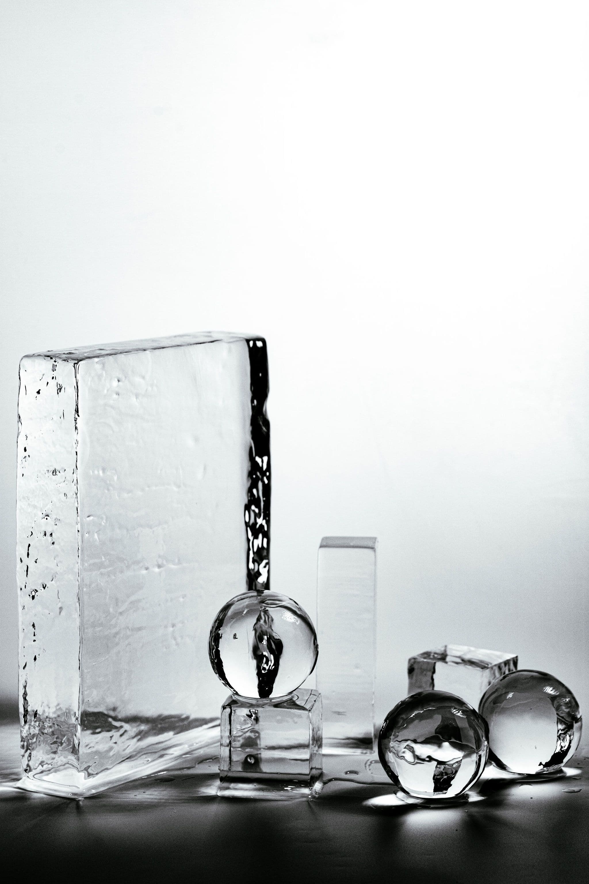 Clear ice blocks, cubes and spheres arranged as a monochrome still life on a white background.