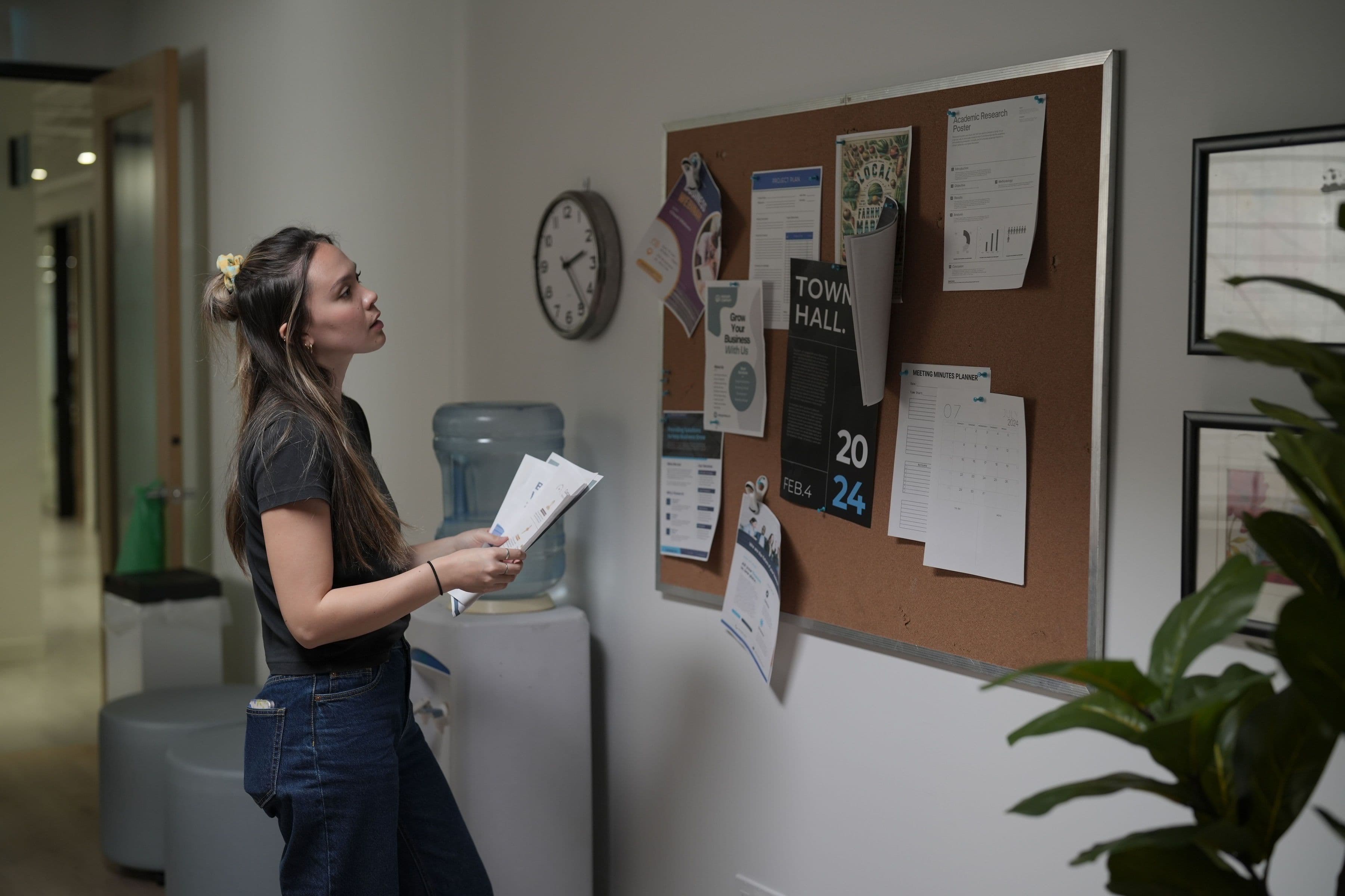 A woman holding papers beside a workplace noticeboard with a Town Hall poster.