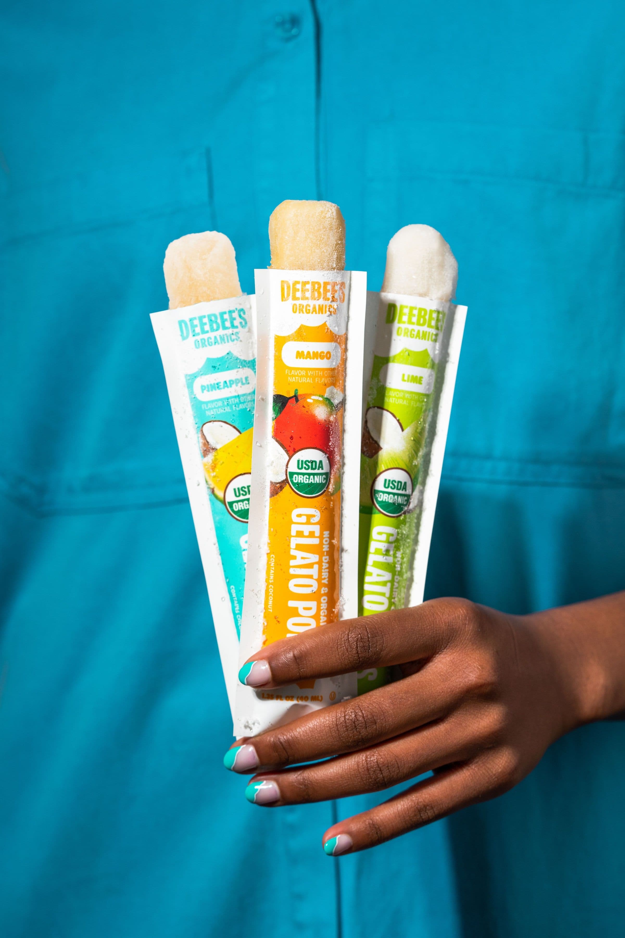 A hand holding three opened DeeBee’s SuperFruit freezies against a turquoise shirt.