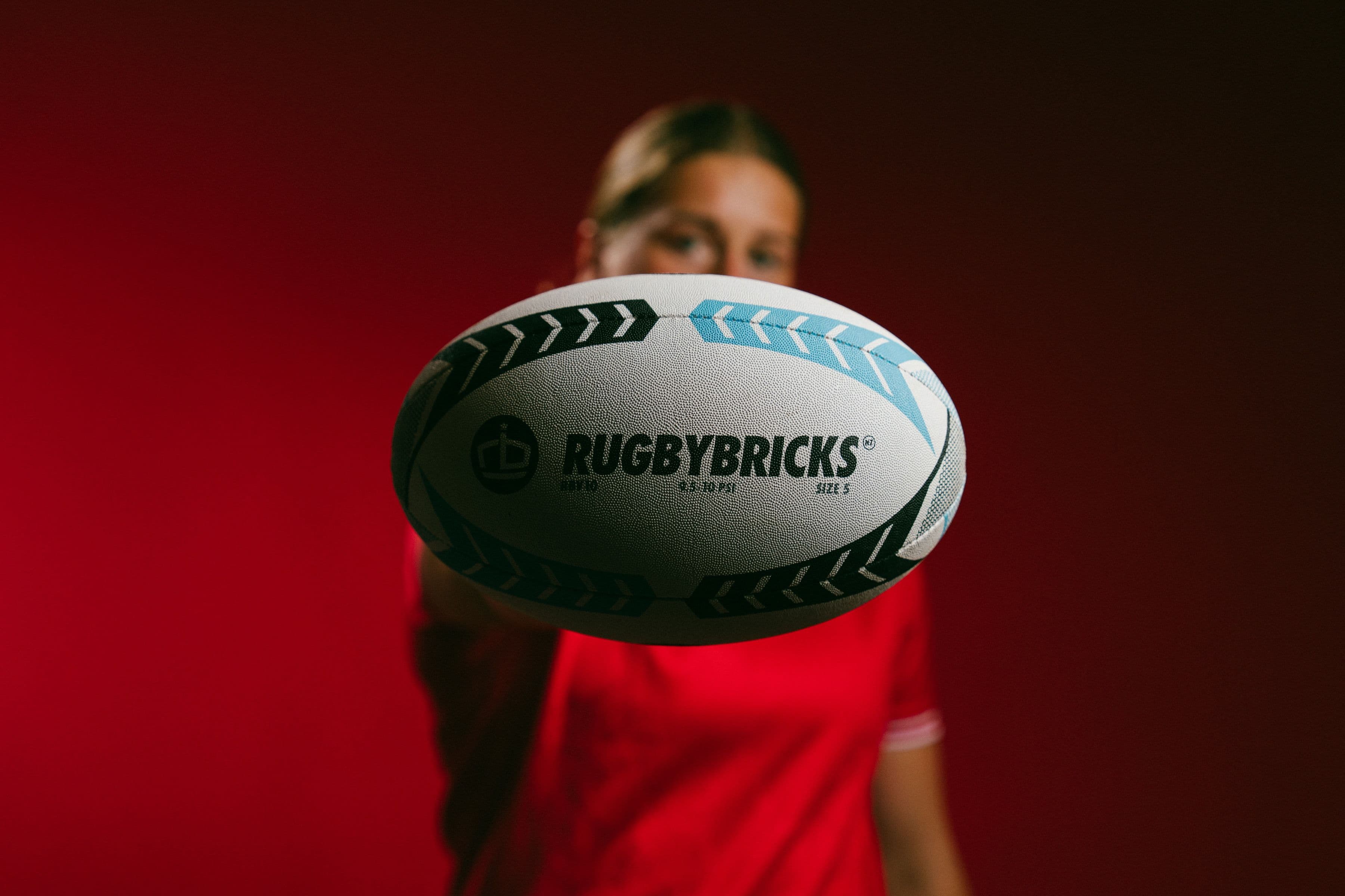 Sophie de Goede holding a Rugby Bricks ball towards the camera against a red studio background.