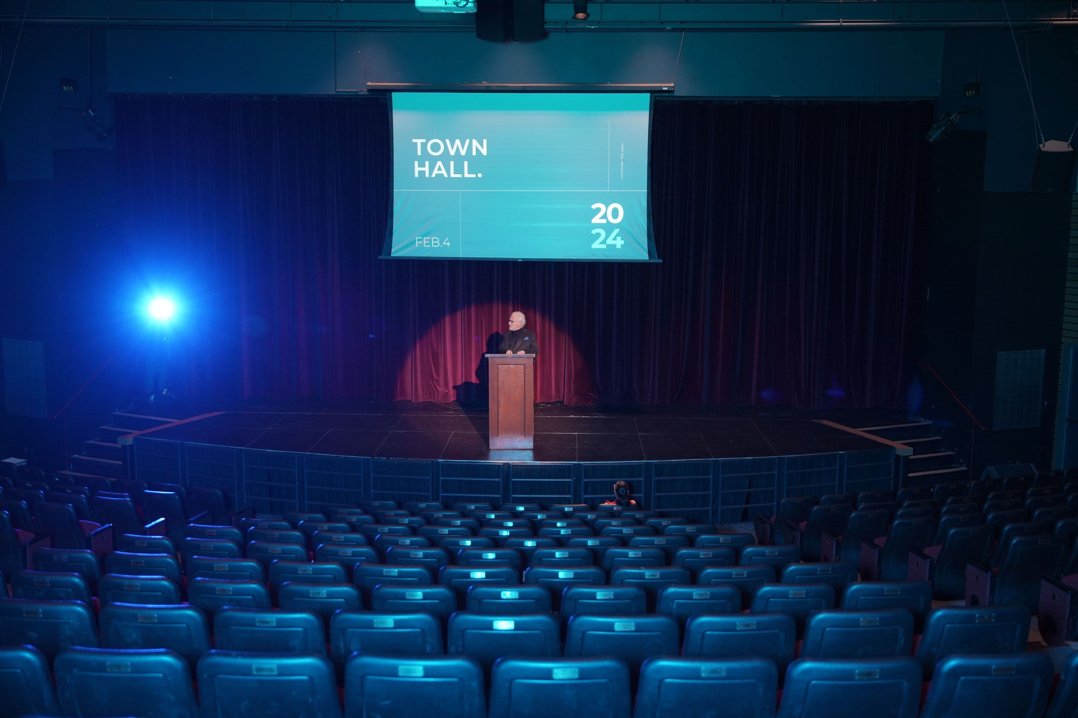 A speaker at a podium on a blue-lit stage facing an empty auditorium, beneath a Town Hall 2024 screen.