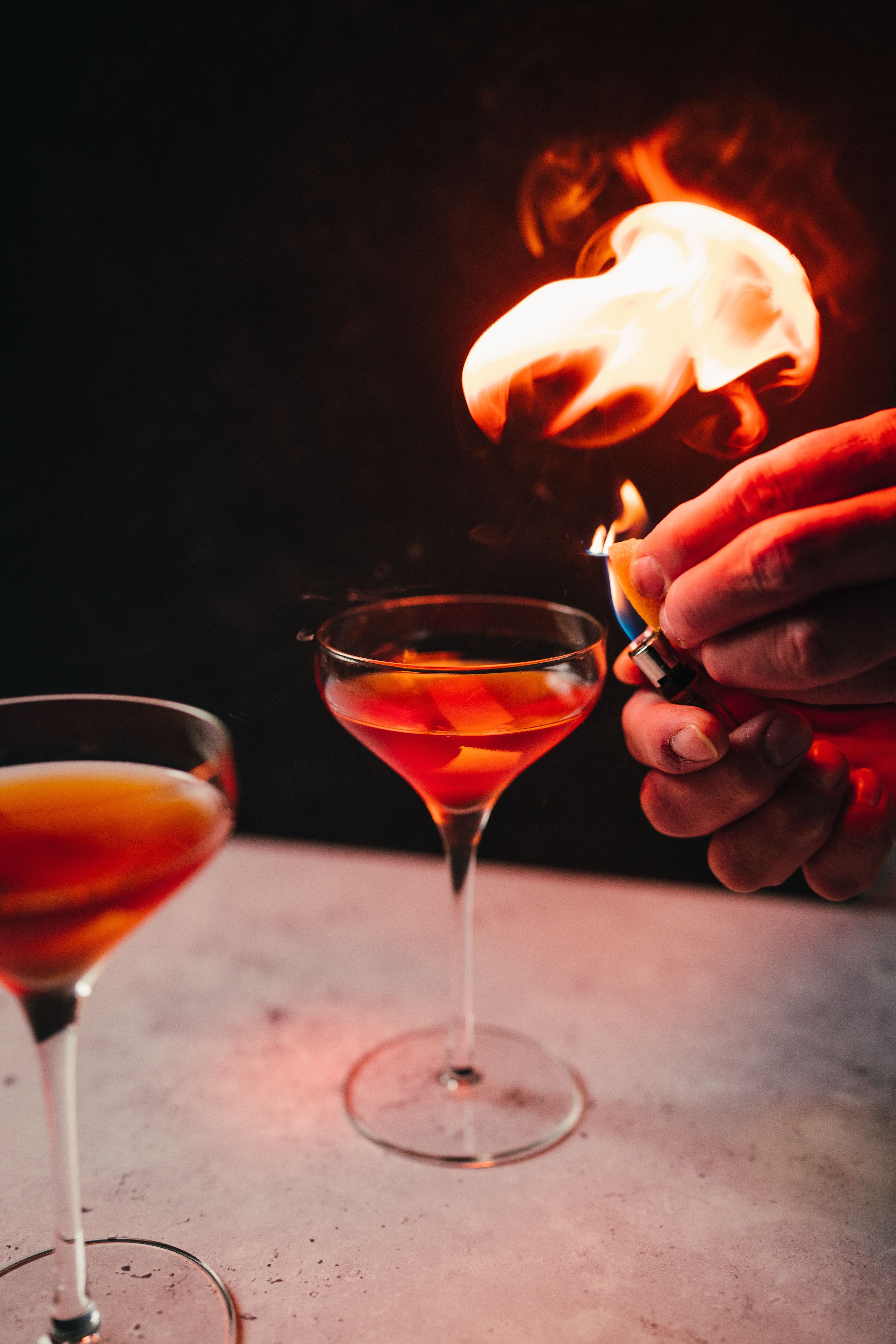 A bartender igniting an orange peel over two amber cocktails in a dark dining room.