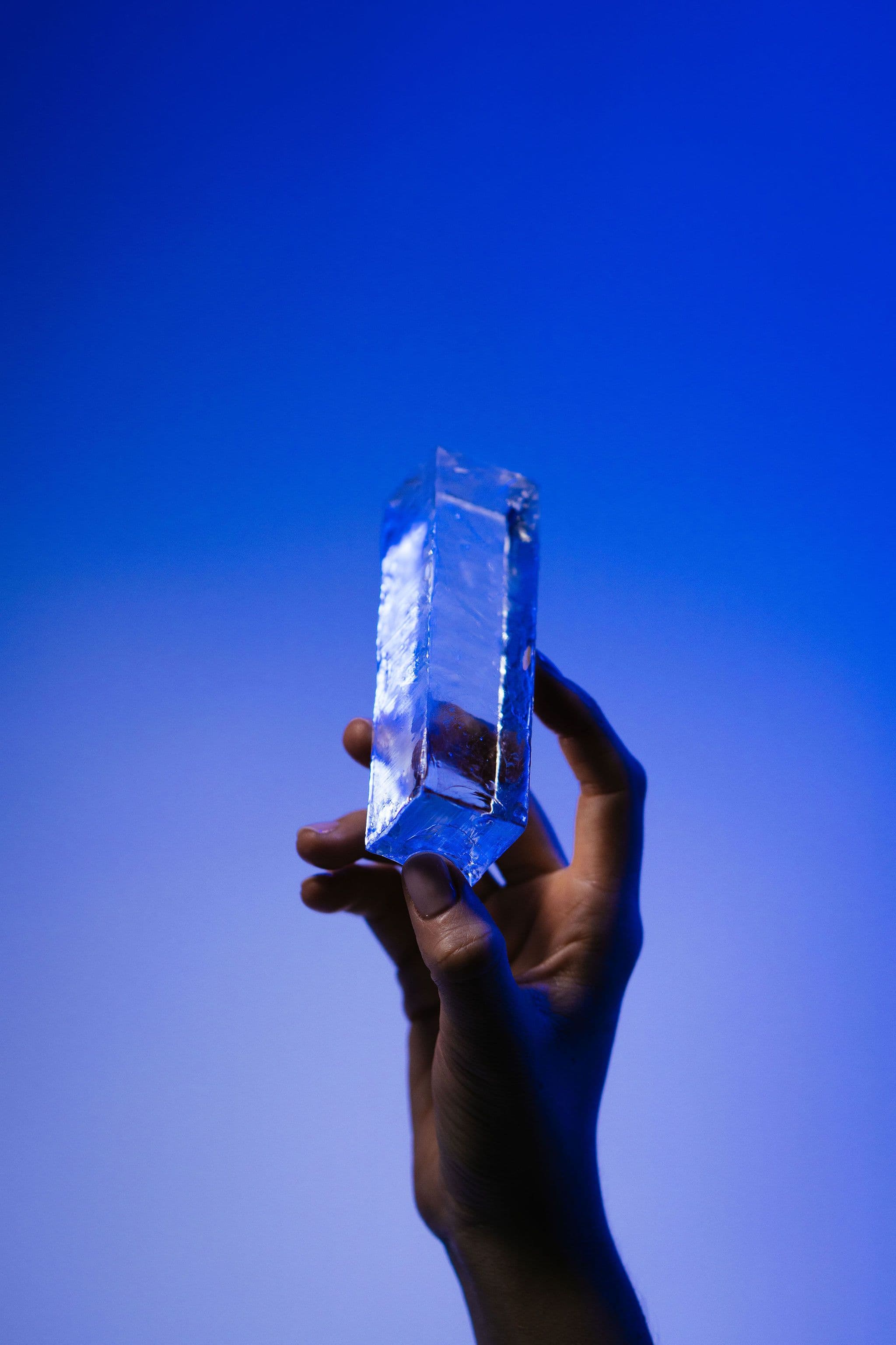 A hand holds a narrow column of clear ice upright against a softly lit blue background.