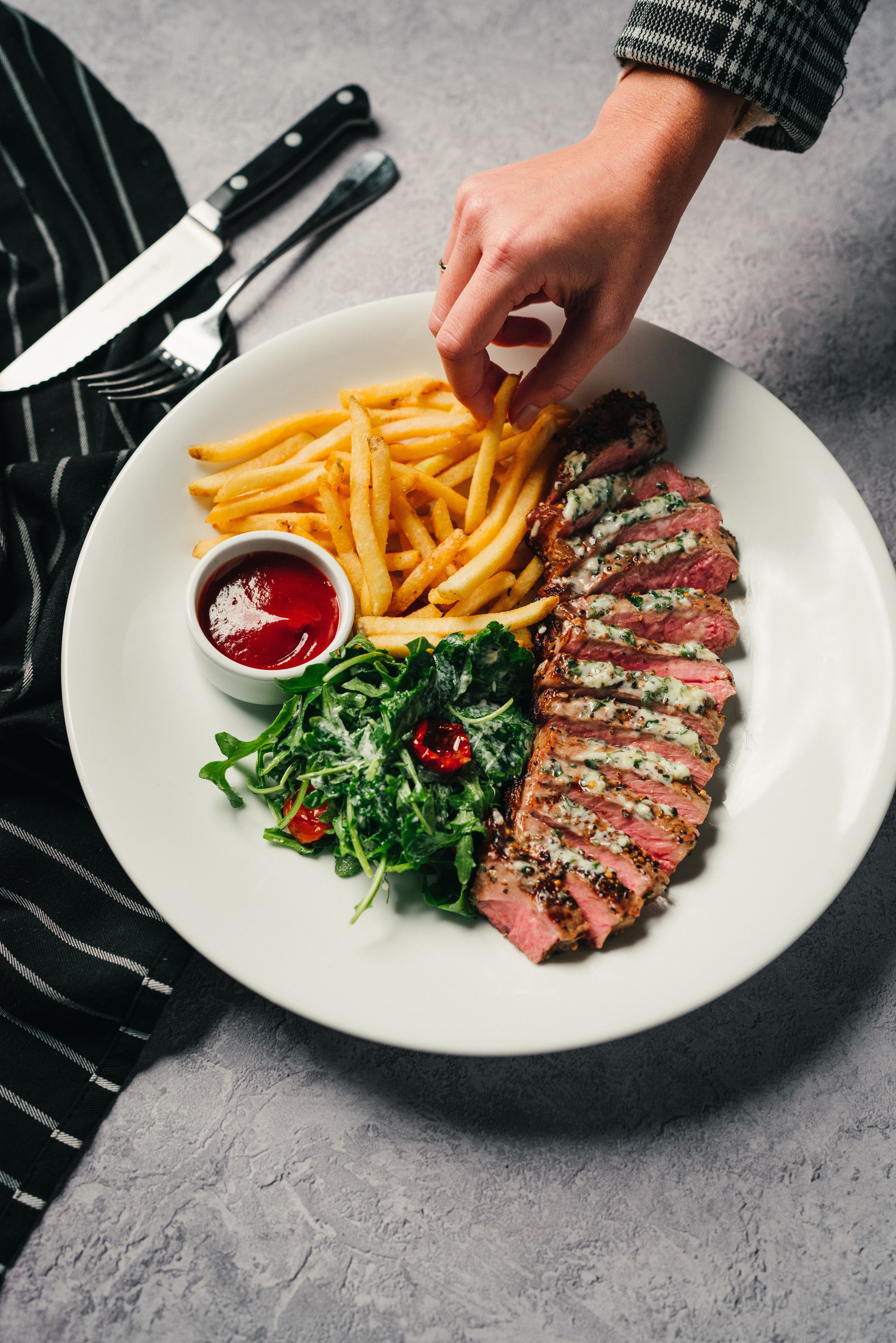 A hand reaching for fries beside sliced steak, greens, and ketchup.
