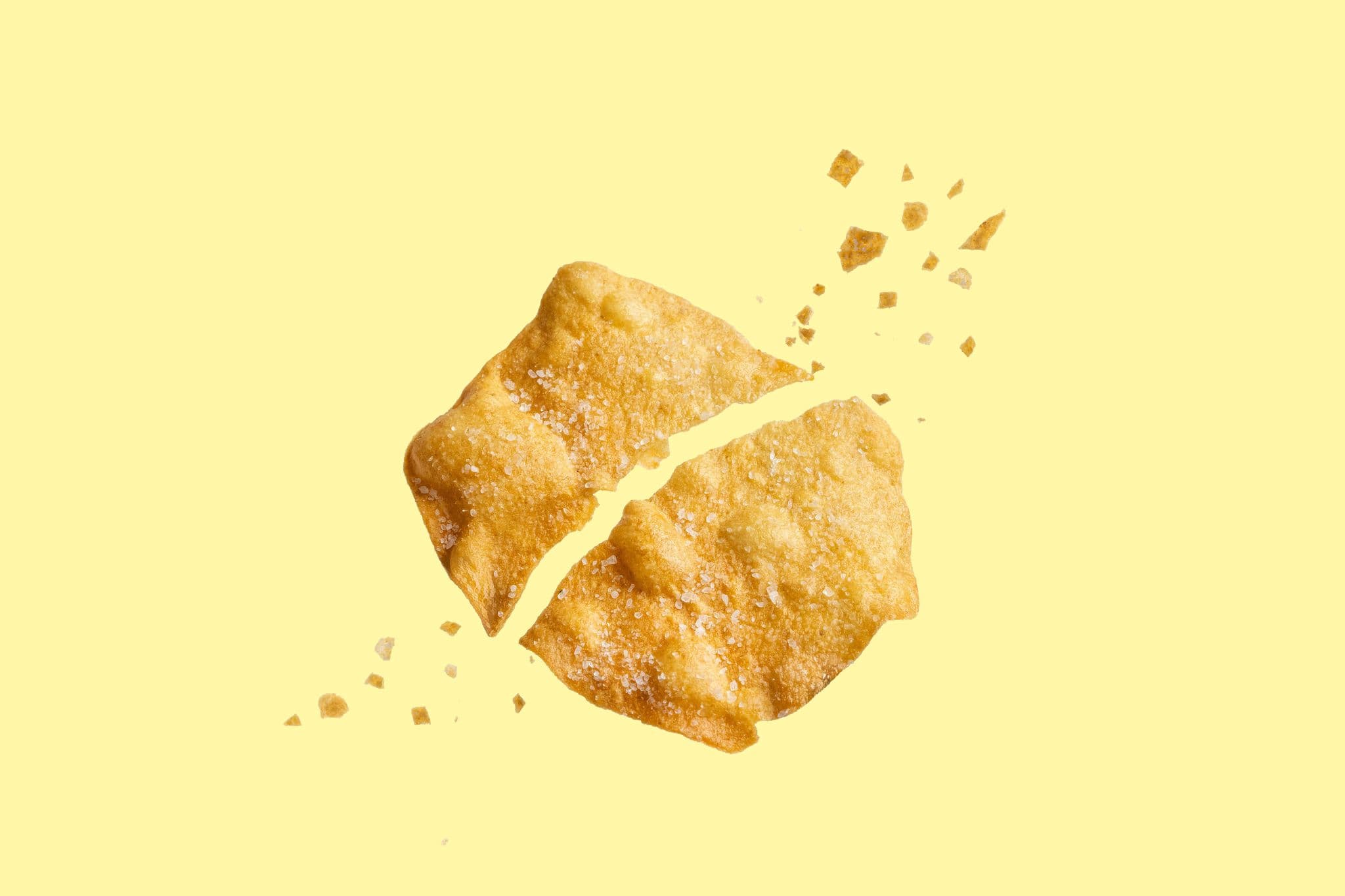 A potato crisp breaking into pieces against a pale yellow background.