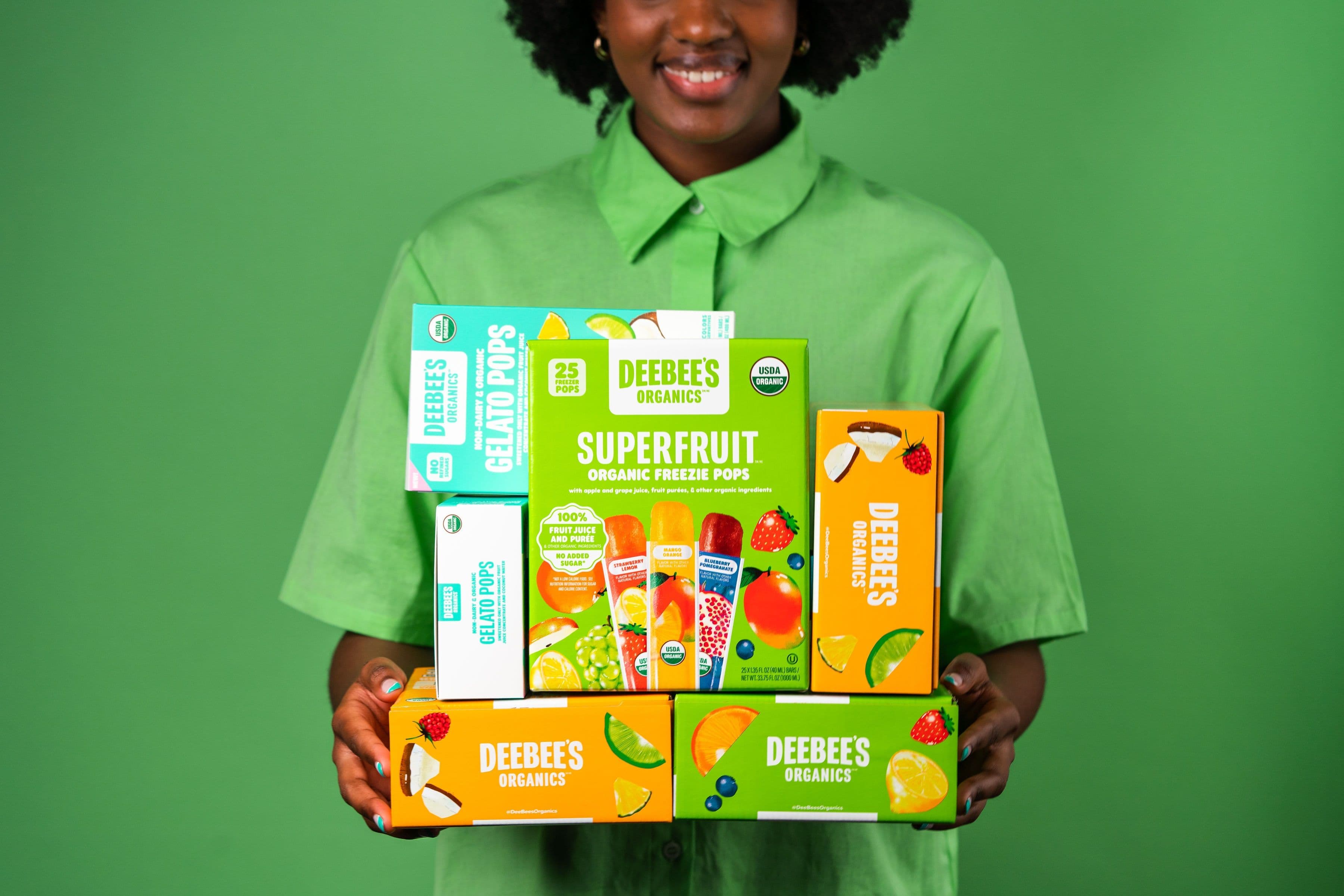 A person in a green shirt holding a stack of colourful DeeBee’s SuperFruit freezer pop boxes against a green background.
