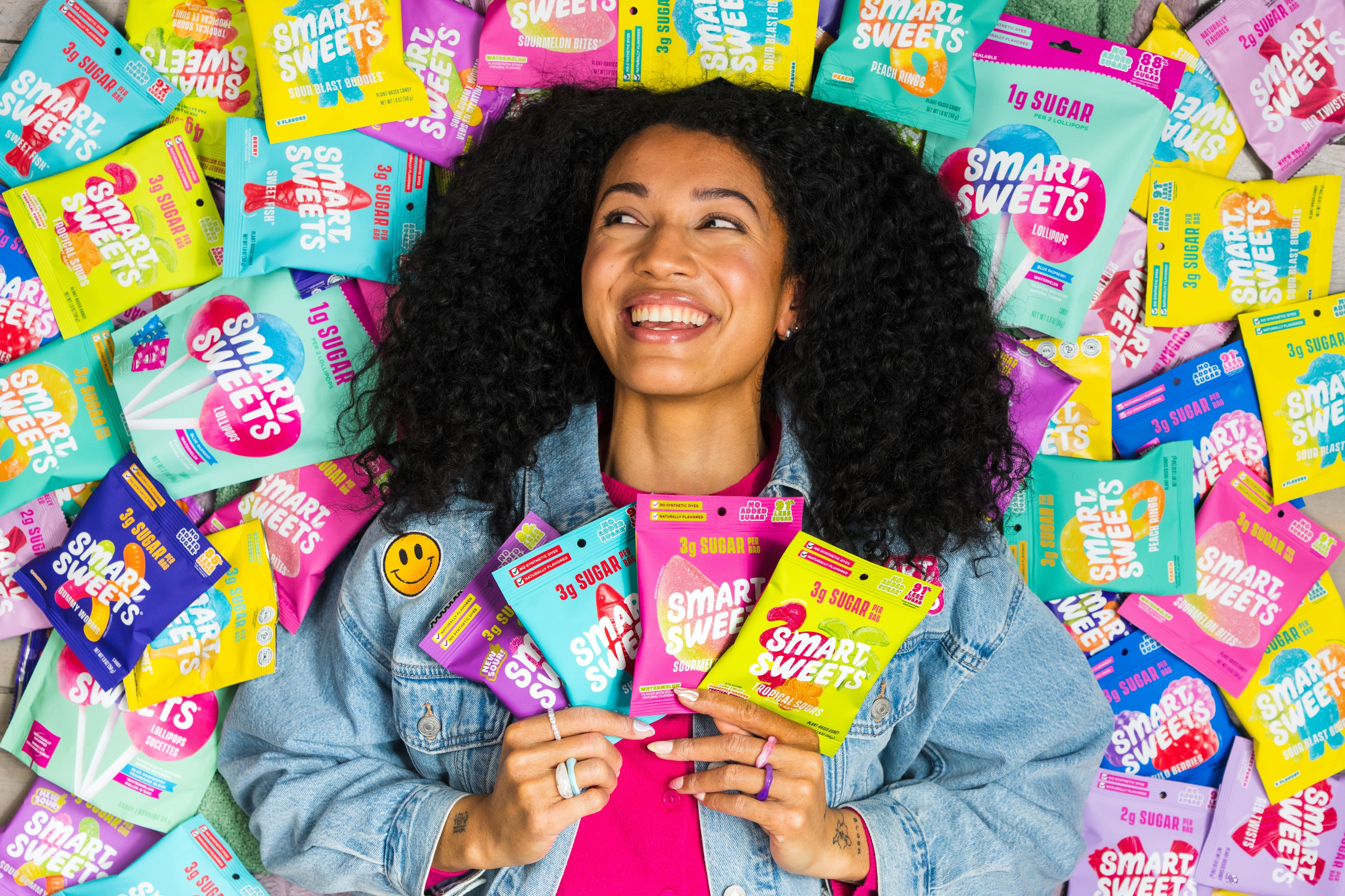 A smiling woman surrounded by a colourful assortment of SmartSweets candy packages.