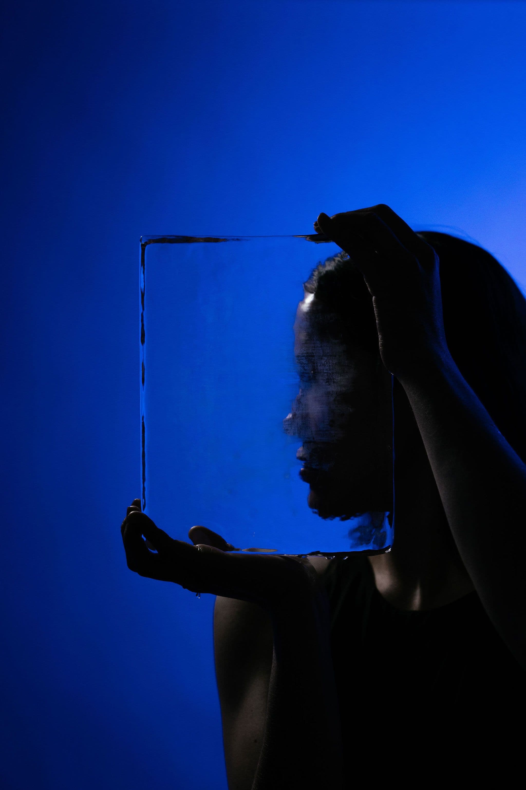 A silhouetted model holds a large rectangular sheet of clear ice in front of her face against a vivid blue background.