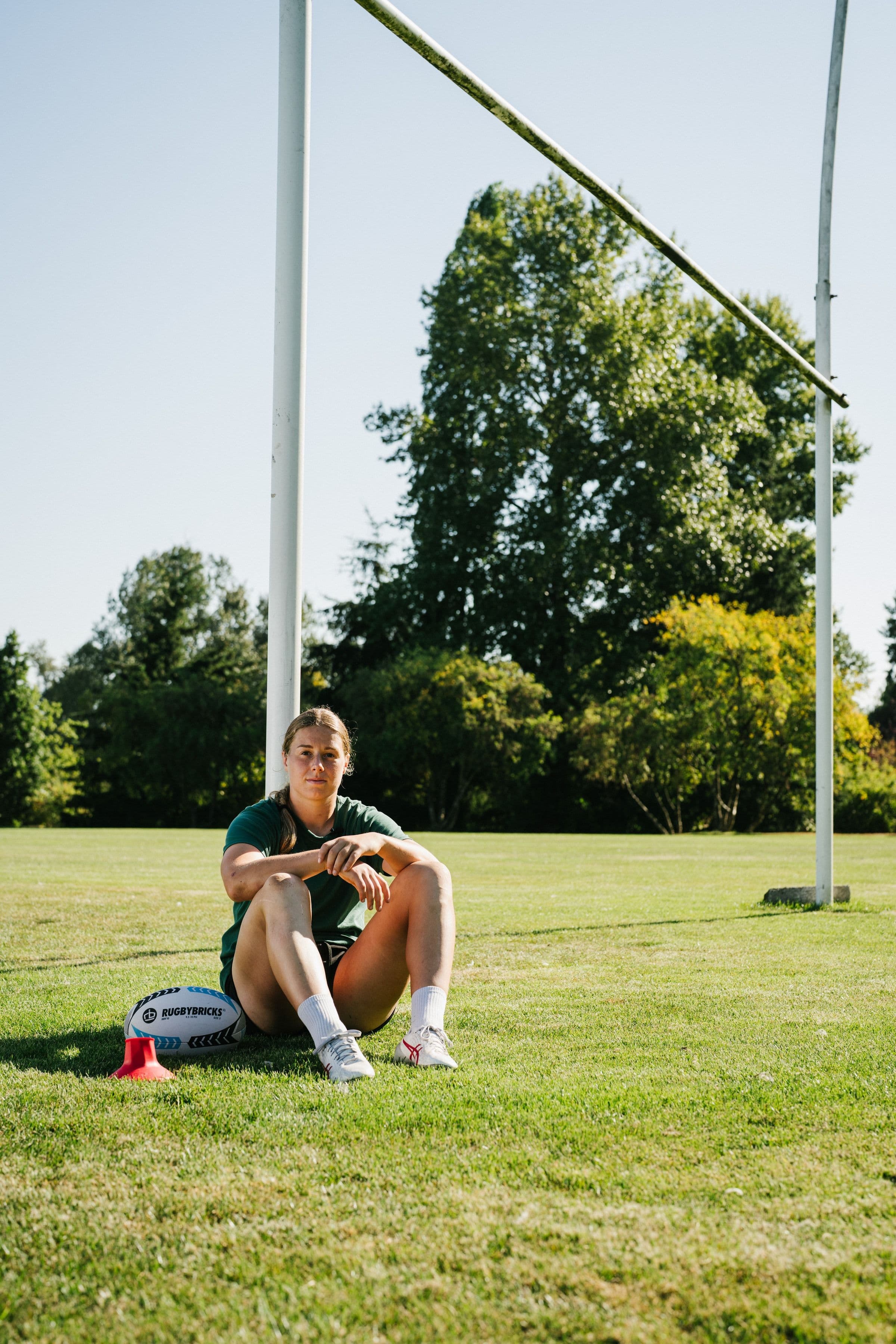 Sophie de Goede seated beneath rugby goalposts beside a Rugby Bricks ball and red kicking tee.
