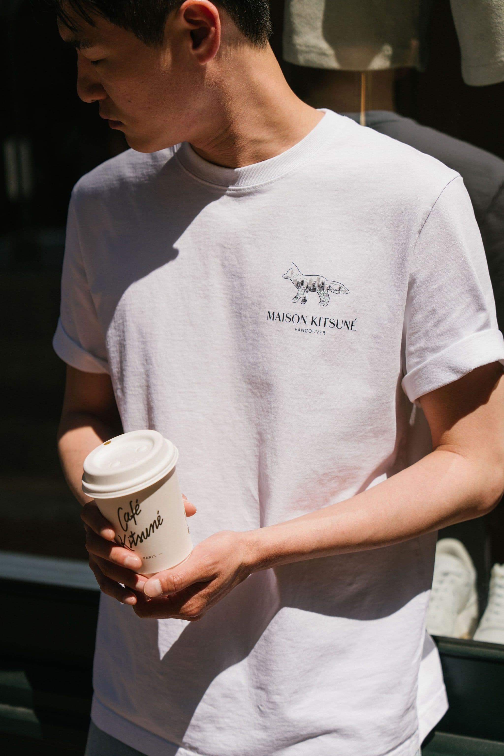 Person wearing a white Maison Kitsuné Vancouver T-shirt and holding a Café Kitsuné takeaway cup.