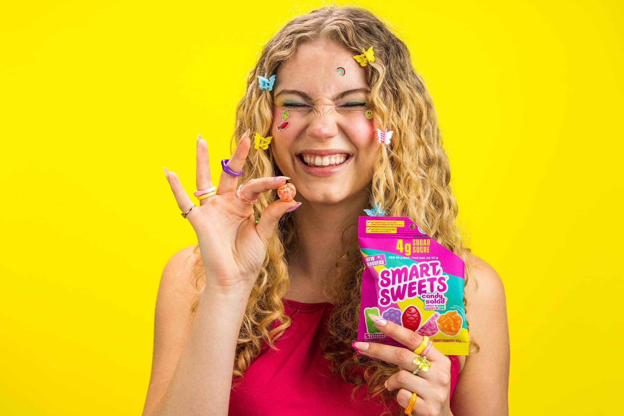 A smiling woman holding SmartSweets candy against a vivid yellow backdrop.