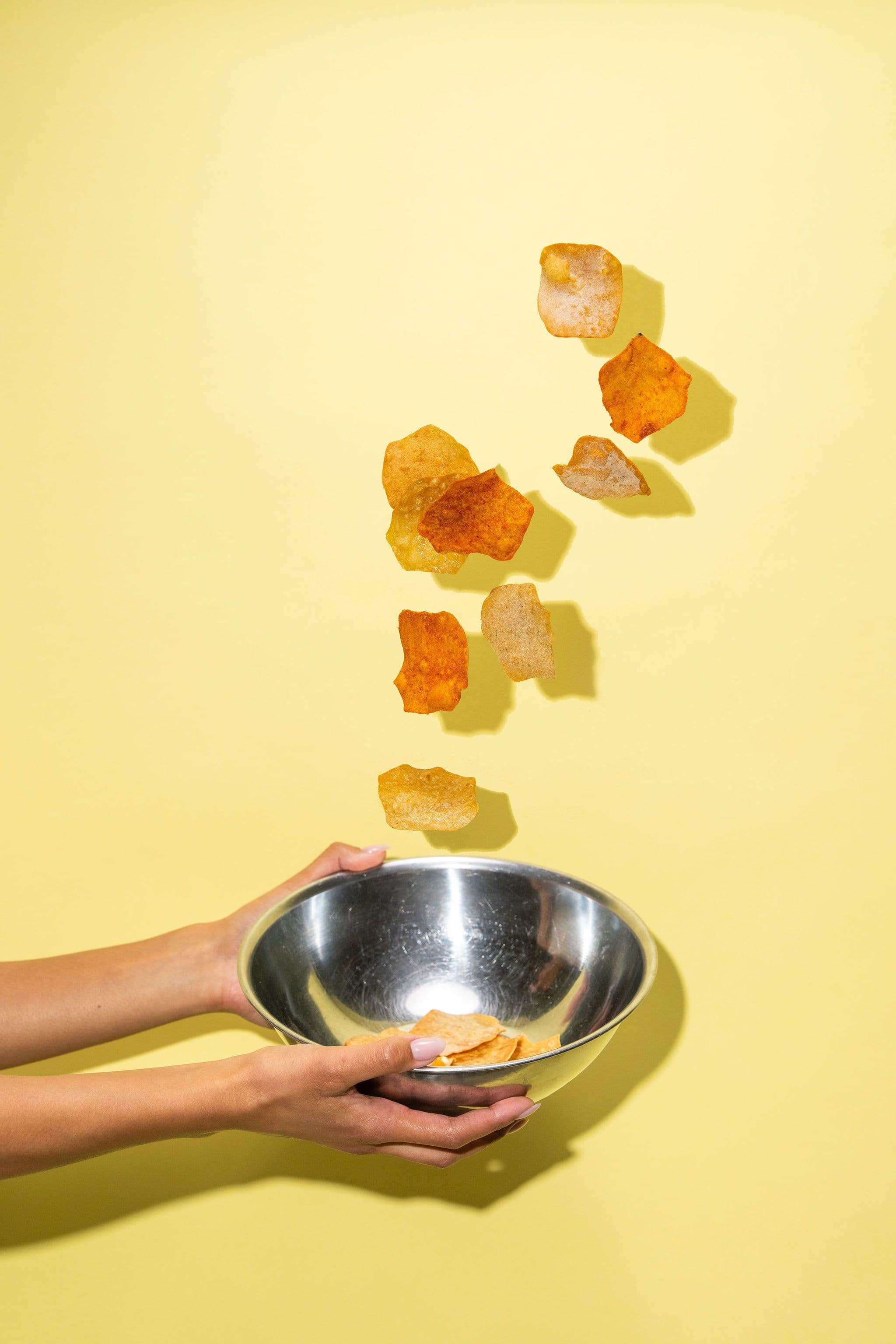 Potato crisps suspended above a metal bowl held against a yellow wall.