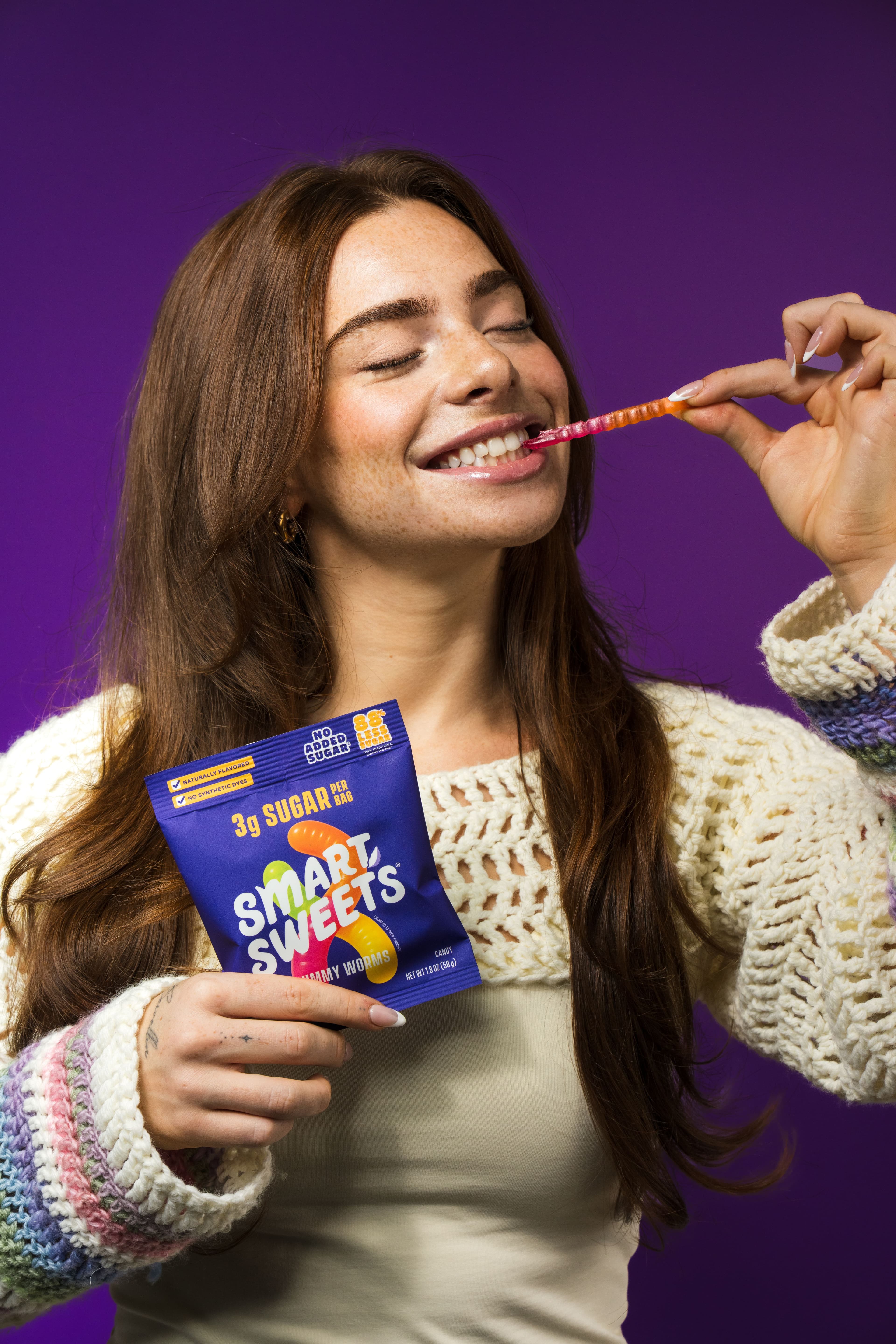 A woman smiling while tasting a SmartSweets sour melon bite against purple.